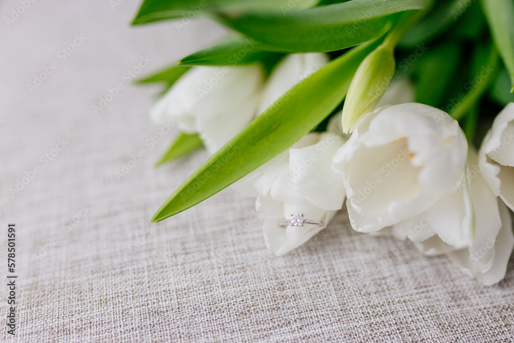 An engagement ring in white gold with a diamond lies in a bouquet of white tulips on white background. Gift for Women's Day, Valentine's Day. Beautiful spring background with green leaves. Mock up An engagement ring in white gold with a diamond lies in a bouquet of white tulips on white background. Gift for Women's Day, Valentine's Day. Beautiful spring background with green leaves. Mock up