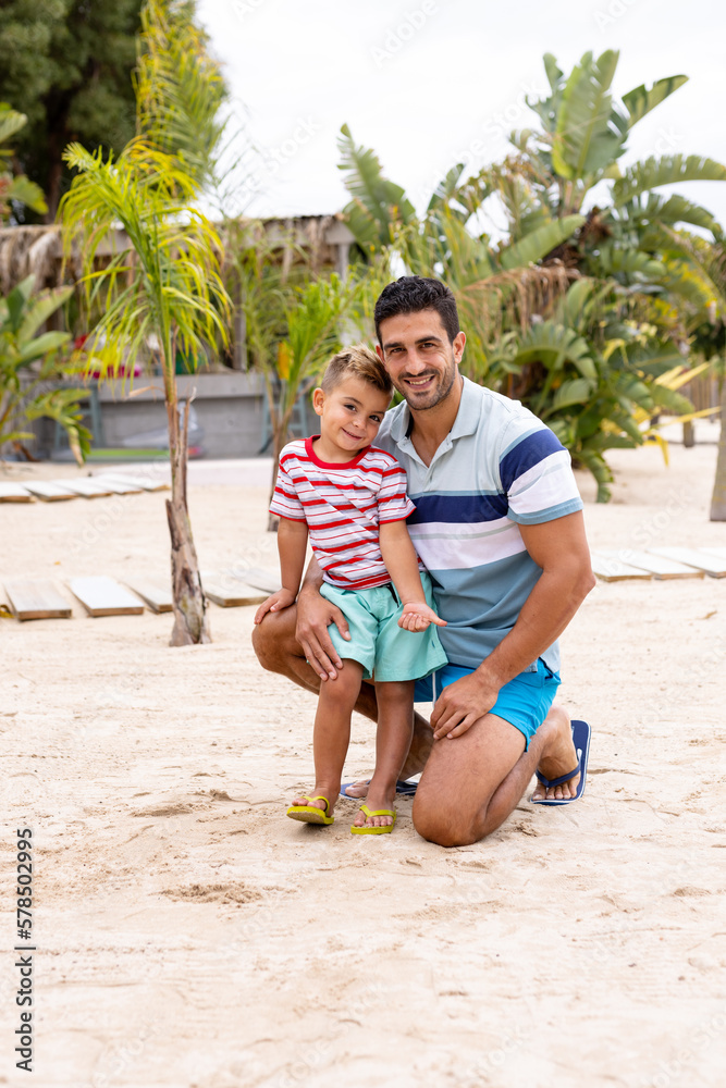 Portrait of happy biracial father and son embracing and smiling at beach, with slow motion - obrazy, fototapety, plakaty 