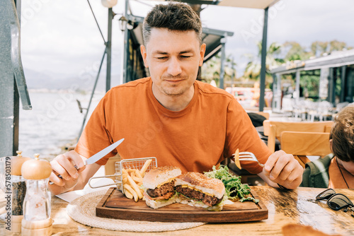 Canvas-taulu Man male with grimace on face looking at fat unhealthy red meat sliced grilled tasty beefy cheese burger hamburger on a plate in the street cafe, while holding fork and knife