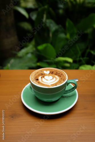 Beautiful green mug with cappuccino or latte coffee on a wooden table, on tropical background. 
