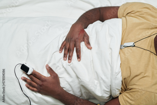 View from above of young sick man with pulse oximeter on fingertip lying under white sheet in hospital bed during intensive care