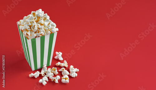 Green white striped carton bucket with tasty cheese popcorn, isolated on red background. Box with scattering of popcorn grains. Fast food, movies, cinema and entertainment concept.