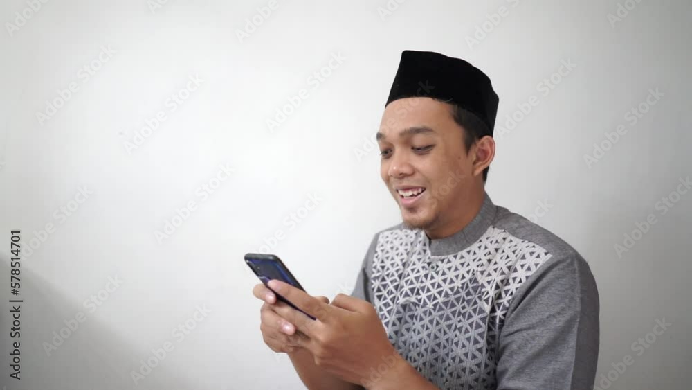 Indonesian Asian muslim man smiling and looking at smartphone wears black hat called peci or songkok