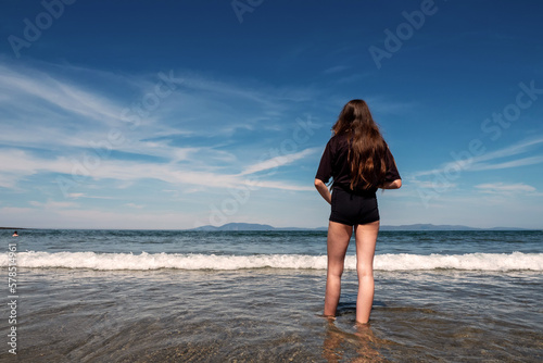 Teenager girl in dark t shirt in the ocean, Blue cloudy sky in the background. Summer time and holiday mood. Enjoy open fresh water and play with waves concept. Irish beach with stunning view.