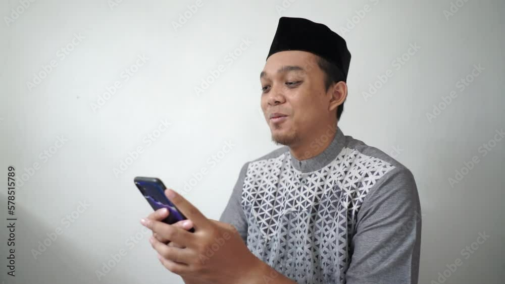Indonesian Asian muslim man smiling and looking at smartphone wears black hat called peci or songkok