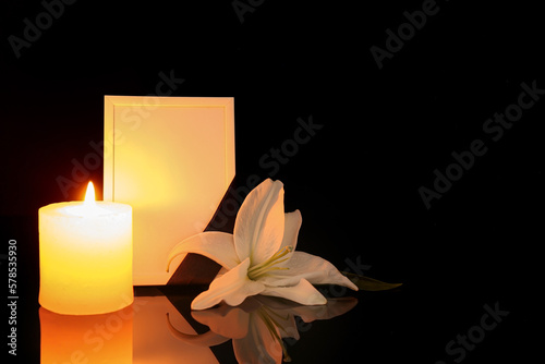 Blank funeral frame, lily flower and burning candle on dark background