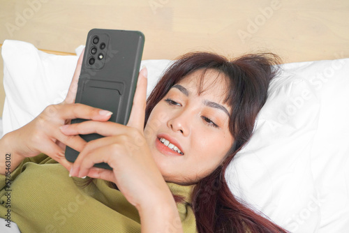 A smiling young Asian woman is laughing while holding her phone and lying on the bed