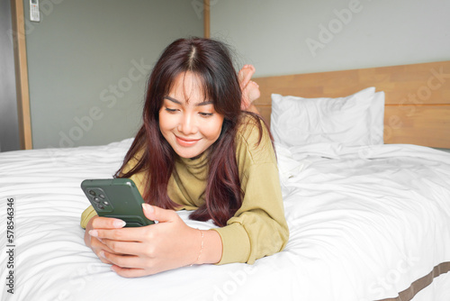 A smiling young Asian woman is laughing while holding her phone and lying on the bed