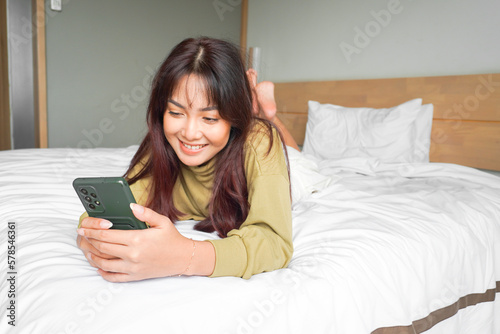 A smiling young Asian woman is laughing while holding her phone and lying on the bed