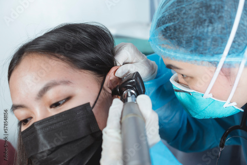 A doctor in full PPE gear checks the ear drum of a female patient with an otoscope for the source of an earache.