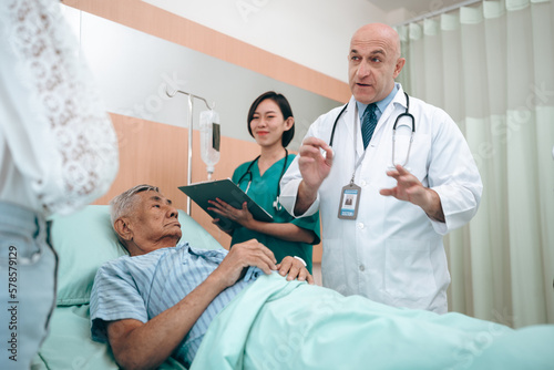 professional doctor standing by explain the treatment information via male elderly patient lying in bed in private hospital sickroom. The treatment program is suitable for people to socialize elderly