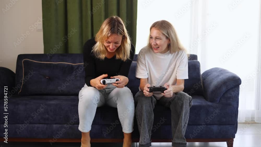 Excited gamer girls sitting on a couch and playing video games, holding joysticks and having fun, woman comes to her friend to play video games together Excited gamer girls sitting on a couch and playing video games, holding joysticks and having fun, woman comes to her friend to play video games together