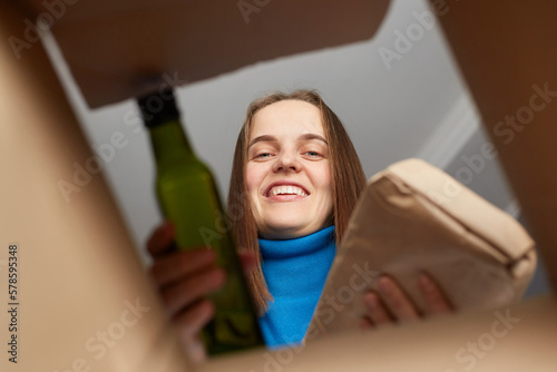 Friendly smiling cute young adult woman looking inside box, putting food donations, grocery help for people in need, groceries for poor humans, humanitarian aid.