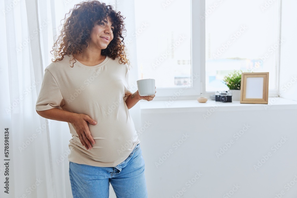 A pregnant woman with curly hair smiling stands at the window with a mug of warm water and tea and enjoys the view in a home t-shirt, happiness of motherhood A pregnant woman with curly hair smiling stands at the window with a mug of warm water and tea and enjoys the view in a home t-shirt, happiness of motherhood