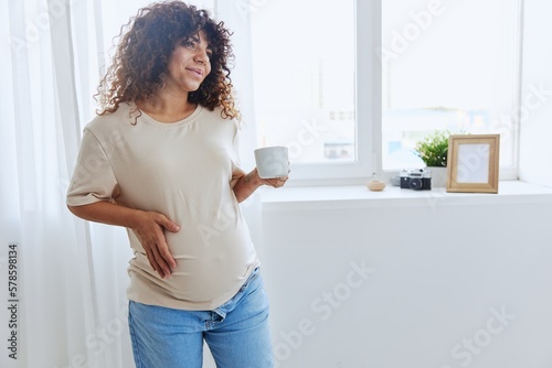 A pregnant woman with curly hair smiling stands at the window with a mug of warm water and tea and enjoys the view in a home t-shirt, happiness of motherhood