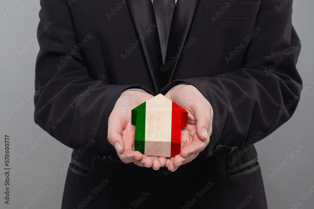 Italian person holding wooden house with flag of Italy Finance, investment, mortgage, loan and credit concept Italian person holding wooden house with flag of Italy Finance, investment, mortgage, loan and credit concept