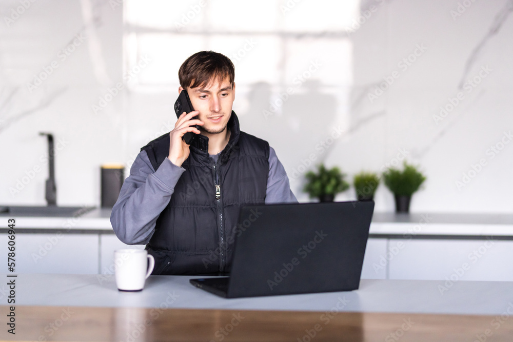 Gig economy concept. Young man freelancer working remotely at home, looking at laptop and consulting client via cellphone, sitting at kitchen Gig economy concept. Young man freelancer working remotely at home, looking at laptop and consulting client via cellphone, sitting at kitchen
