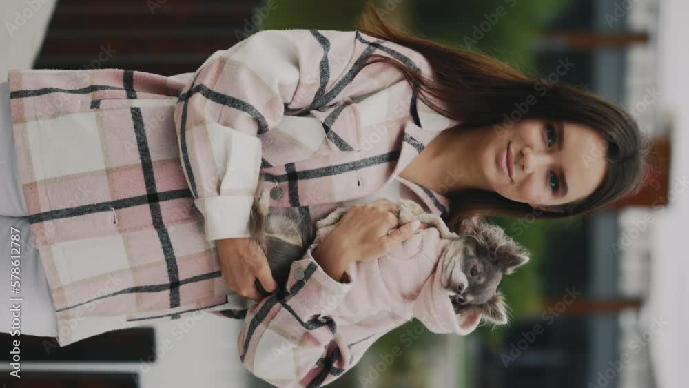 Vertical Video of the Beautiful Caucasian Woman holding the Chihuahua wearing Stylish Dog Clothes, standing looking at the camera on the blurred background. Portrait of Pretty Girl with her Pet Vertical Video of the Beautiful Caucasian Woman holding the Chihuahua wearing Stylish Dog Clothes, standing looking at the camera on the blurred background. Portrait of Pretty Girl with her Pet