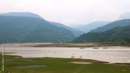 Wooden boats glide on a river in Bholaganj, Sylhet, Bangladesh-India border, as fishermen work beneath clouds on a hill. photo