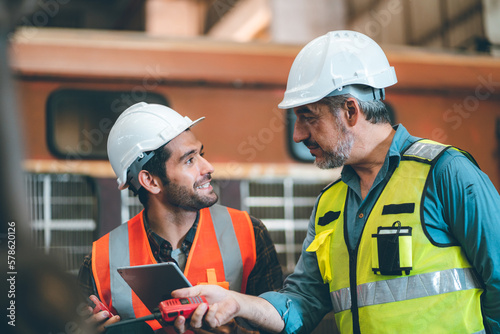 Two senior and young male Asian engineers in safety vest and jacket with hardhat and helmet working in warehouse and factory on a machine while fixing and inspecting equipment