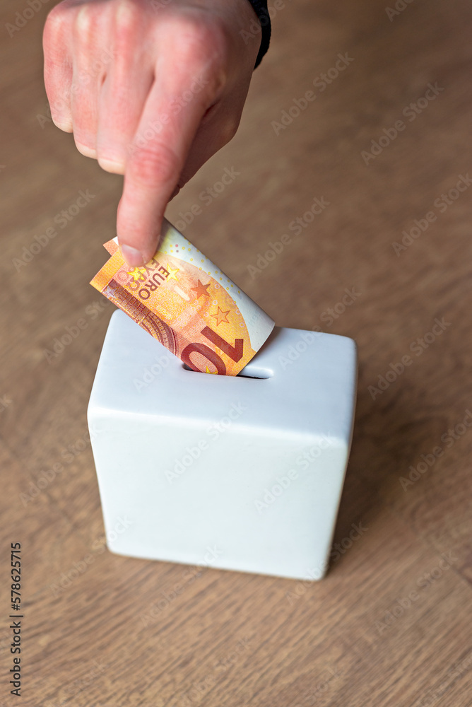 Putting a euro bill into a white square piggy bank. Text space.