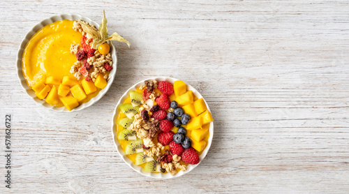 Ywo Healthy mango smoothie bowls with blueberries, raspberries, kiwi and granola. Above view scene on a light grey  wooden background.