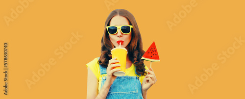 Summer colorful portrait of young woman drinking fresh juice with lollipop or ice cream shaped slice of watermelon wearing sunglasses on orange background
