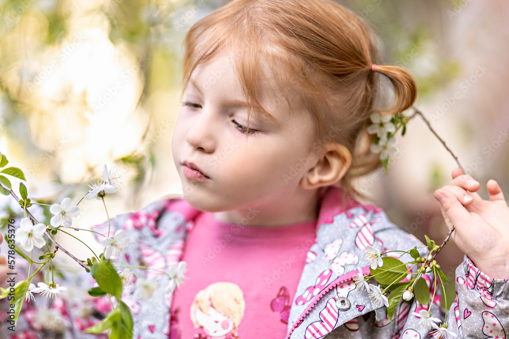 Fototapeta premium Close-up portrait of a toddler girl with red hair in front of a cherry blossom. Spring