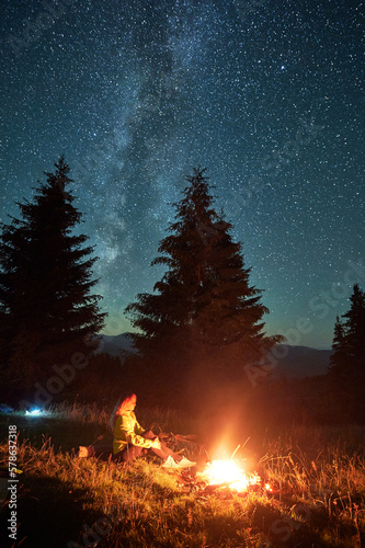 Night camping in mountains under starry sky with Milky way. Woman sitting on grass near campfire, admiring landscape, resting, enjoying. Concept of harmony with nature.