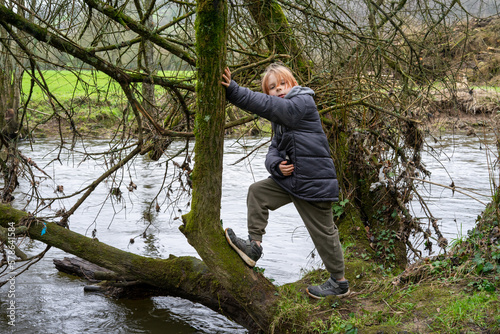 A young boy on a fallen tree near the river. A boy with long hair in a warm jacket is nine years old.
