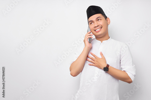 Friendly smiling young Asian Muslim man in Arabic clothes talking on mobile phone with hand his chest with grateful expression isolated on white background. People religious Islamic lifestyle concept