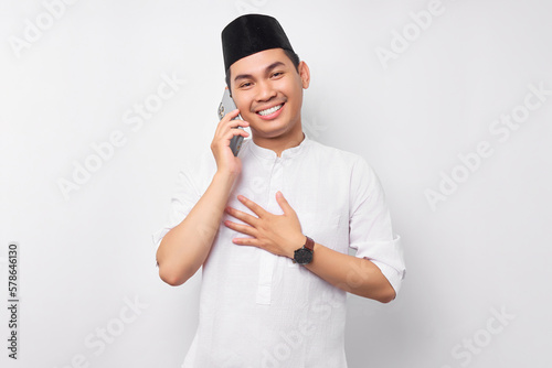 Friendly smiling young Asian Muslim man in Arabic clothes talking on mobile phone with hand his chest with grateful expression isolated on white background. People religious Islamic lifestyle concept