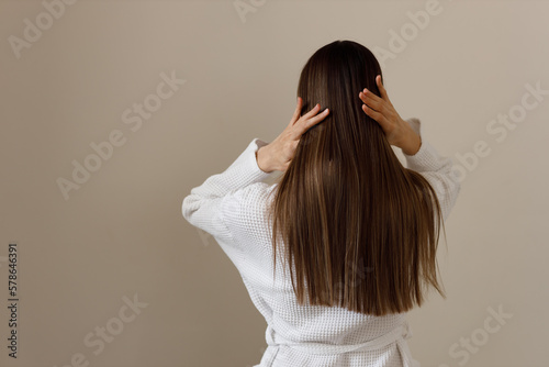 Young woman with healthy long smooth hair in white bathrobe, rear view. Hygiene, freshness, cleansing after shower. Effect of cosmetic products for hair and body care. Healthy cosmetic hair care.