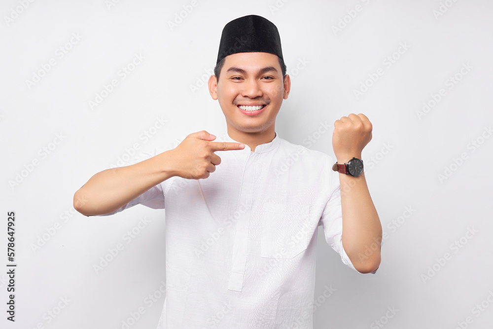 Smiling young Asian Muslim man in Arabic clothes pointing a finger at his wristwatch, indicate the time of the breaking fast isolated on white background. People religious Islamic lifestyle concept Smiling young Asian Muslim man in Arabic clothes pointing a finger at his wristwatch, indicate the time of the breaking fast isolated on white background. People religious Islamic lifestyle concept