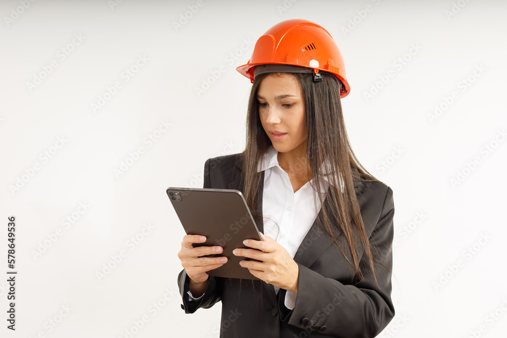 Portrait of young woman with tablet in her hands, dressed in business clothes on isolated white background. Studio shot of attractive happy brunette girl in orange construction helmet on her head. Portrait of young woman with tablet in her hands, dressed in business clothes on isolated white background. Studio shot of attractive happy brunette girl in orange construction helmet on her head.