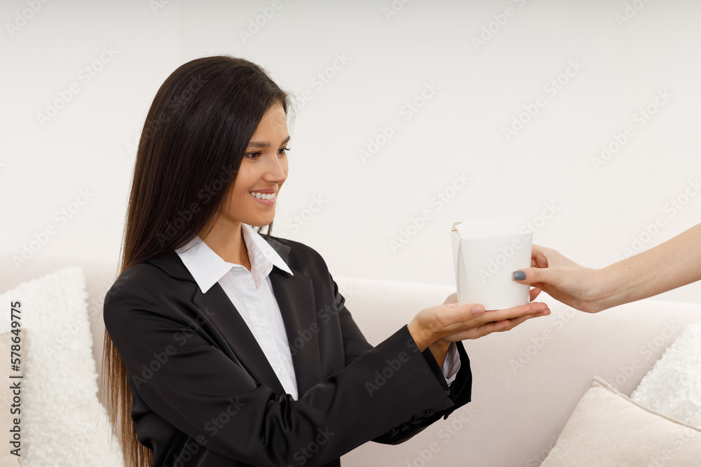 Happy pretty young woman in formal suit and white shirt, sitting on the sofa, stretches out her hands to receive a paper box with food. Fast food during the lunch break in the office. Food delivery. Happy pretty young woman in formal suit and white shirt, sitting on the sofa, stretches out her hands to receive a paper box with food. Fast food during the lunch break in the office. Food delivery.