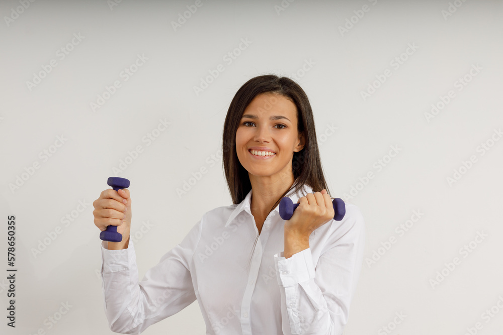 Attractive girl with dark hair in stylish business suit and white shirt holds small female dumbbells. Portrait of happy smiling young woman with sports equipment on isolated white background. Attractive girl with dark hair in stylish business suit and white shirt holds small female dumbbells. Portrait of happy smiling young woman with sports equipment on isolated white background.