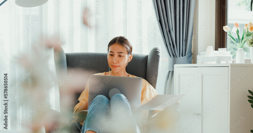 Φωτογραφία Young asia female girl or university student sit on sofa chair with computer laptop wear earphone hold paperwork online meeting presenting discuss work in living room at house