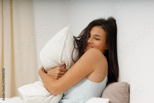 Beautiful slender young woman, dressed in gently light blue silk nightie negligee, lies in cozy bed in the bedroom. Attractive girl hugs pillow after waking up from sleep. Good morning.