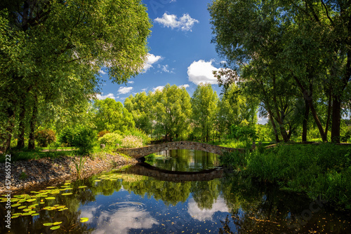 Fototapeta Naklejka Na Ścianę i Meble -  Nature park in summer with river, stone bridge, green trees, and blue sky with white clouds