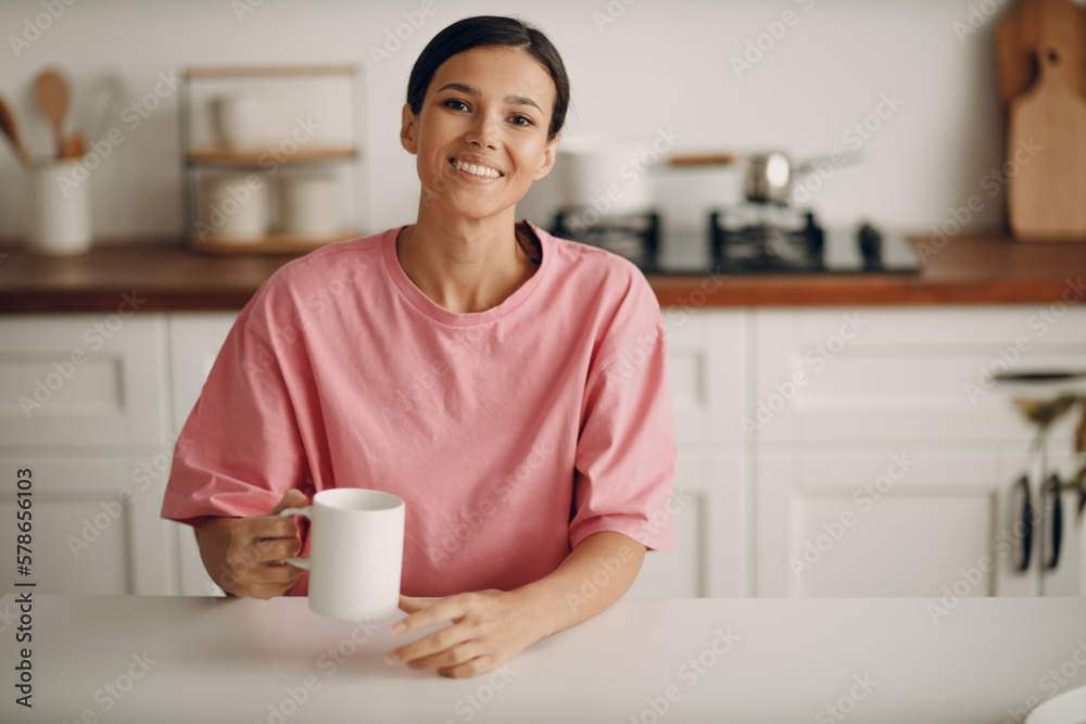 Cute young brunette woman in casual clothes looks straight into the camera. Portrait of smiling happy girl sitting at home in the kitchen at the dining table. Diet, fasting. Cute young brunette woman in casual clothes looks straight into the camera. Portrait of smiling happy girl sitting at home in the kitchen at the dining table. Diet, fasting.