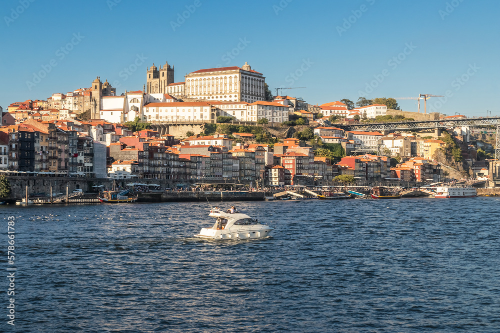 View over Porto from Vila Nova de Gaia, with the Douro River in the foreground and the riverside pier, the houses and the Cathedral in the background, on a summer afternoon.