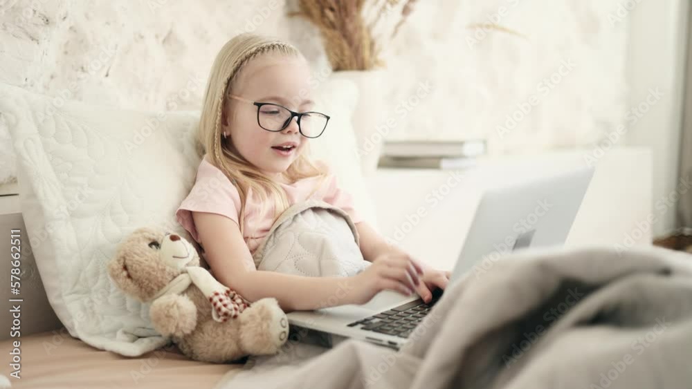Portrait of cute little blond girl kid has video call while laying on a bed in light living room Adorable smiling child talking on video conference online distance remote education at home Portrait of cute little blond girl kid has video call while laying on a bed in light living room Adorable smiling child talking on video conference online distance remote education at home