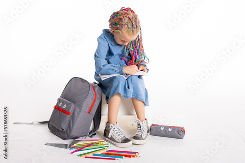 Happy stylish cute adorable school girl sitting on white background islakted holding pencils and notebook doing home assigment homework while shooting process. photo