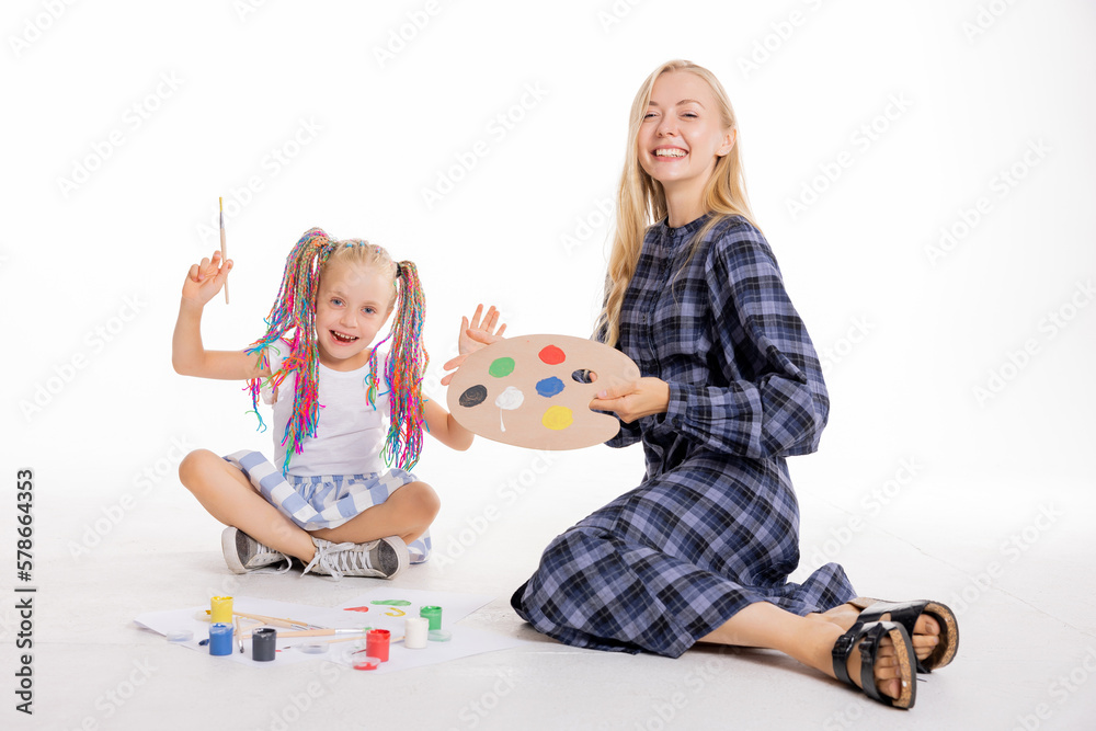 Curious school girl with colorful braids sitting on floor in white studio isolated looking at camera spends time with young mom painting using different art tools. Happy family concept. Curious school girl with colorful braids sitting on floor in white studio isolated looking at camera spends time with young mom painting using different art tools. Happy family concept.