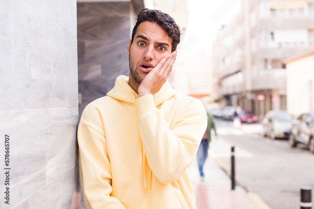 young hispanic man feeling shocked and astonished holding face to hand in disbelief with mouth wide open