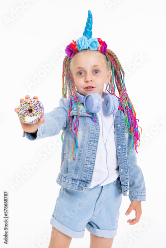 Adorable little girl with colorful barids and unicorn headband with flowers and blue corn standing on white background wearing denim clothes and stylish blue headphones showing donut in hand. photo