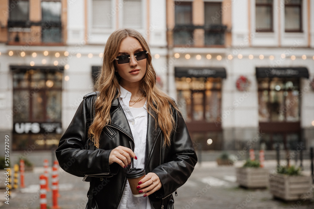 Fashionable  blonde woman model with  black leather jacket, style sunglasses  and coffee walking the city street