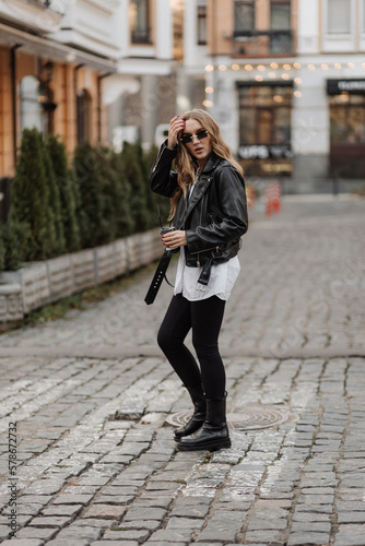 Fashionable blonde woman model with black leather jacket, style sunglasses and coffee walking the city street