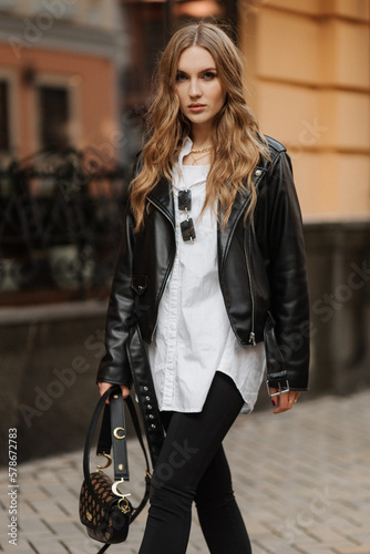 Fashionable blonde woman model with black leather jacket and style bag walking the city street